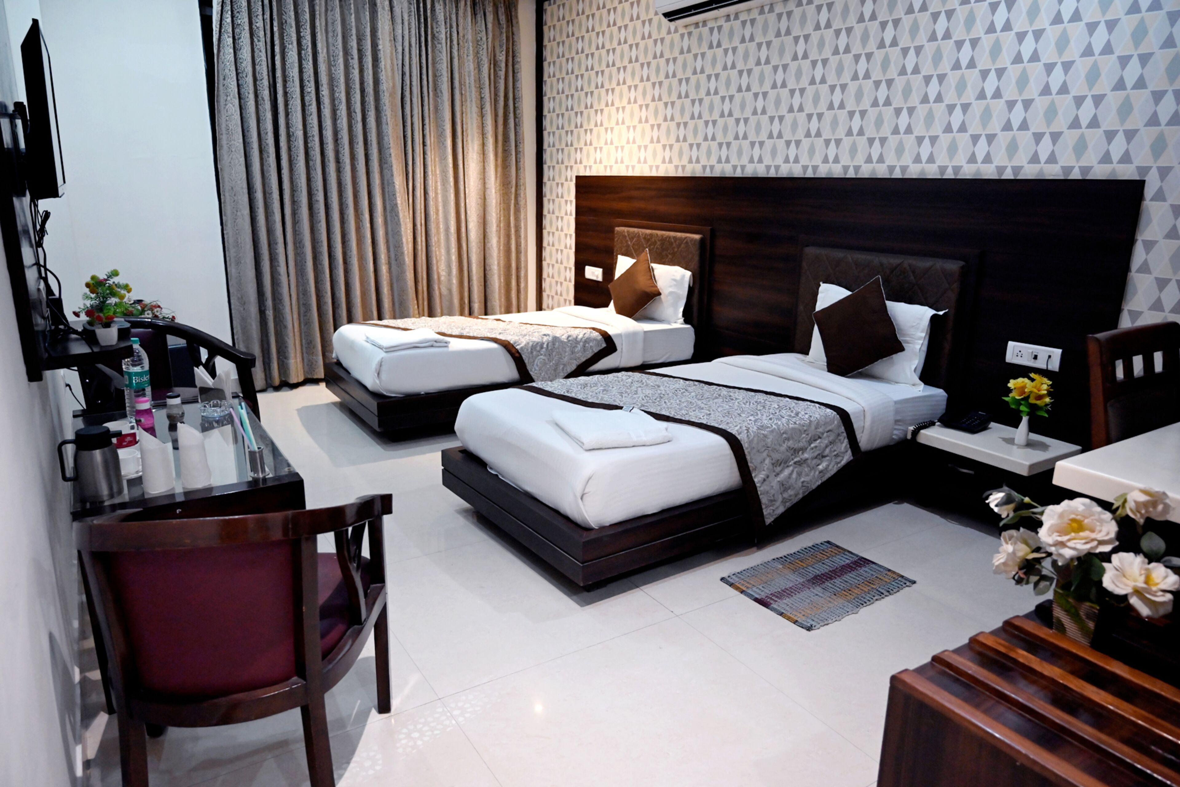 A hotel room with two beds, a desk, and a chair. There is a TV on the wall and a window with curtains.