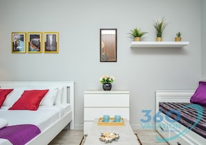 A modern bedroom with a white bed, red and purple pillows, a coffee table with decorative items, a white nightstand, and a wall-mounted shelf with plants. - Stylish Studio in Dubai Marina Close to the Beach, Mall and Metro (Dubai)