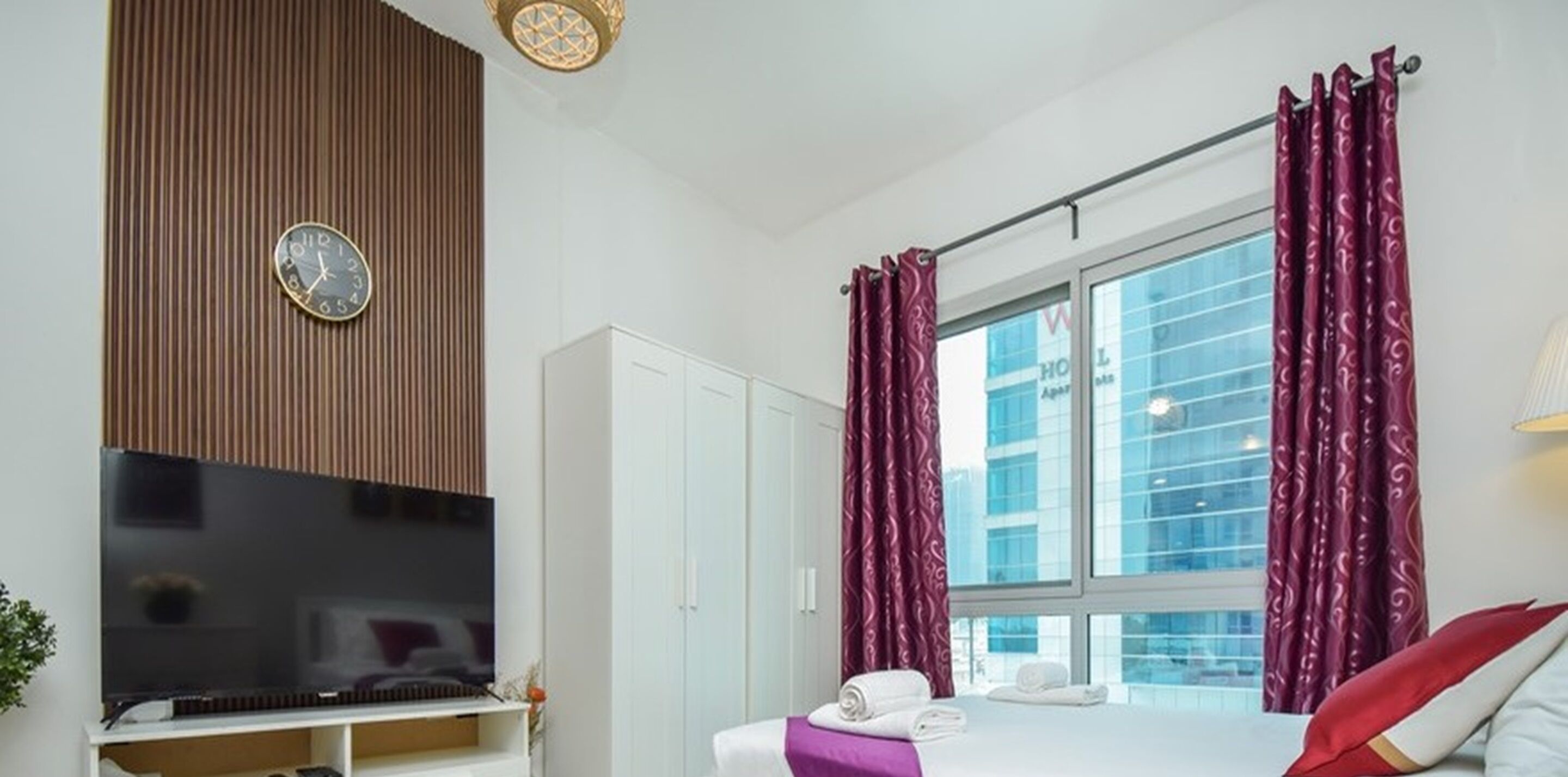 A modern hotel room with a flat-screen TV, a large window with curtains, a bed with pillows, and a wardrobe.