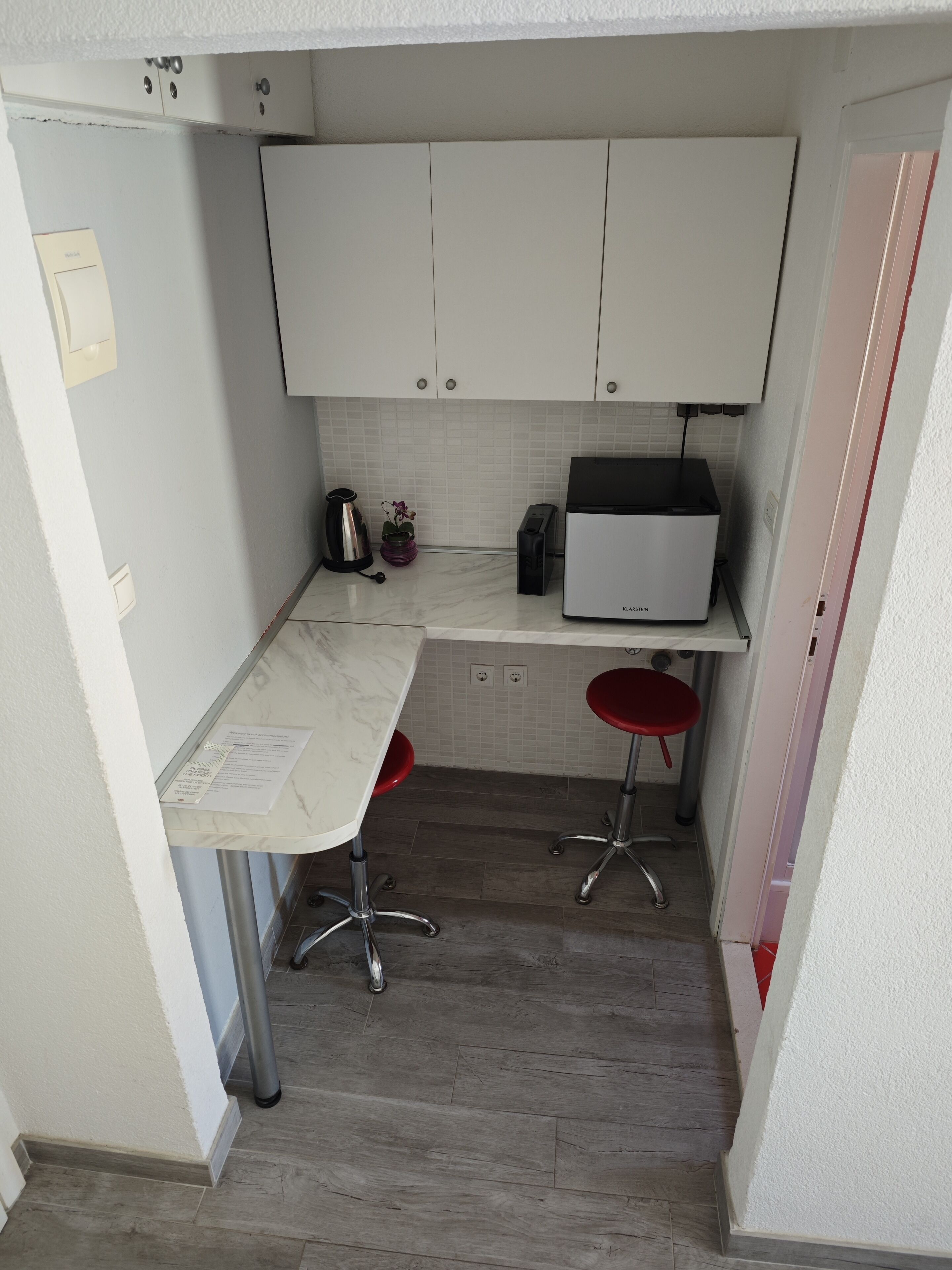 A compact kitchen with white cabinets, a countertop microwave, a small sink, and two red stools.