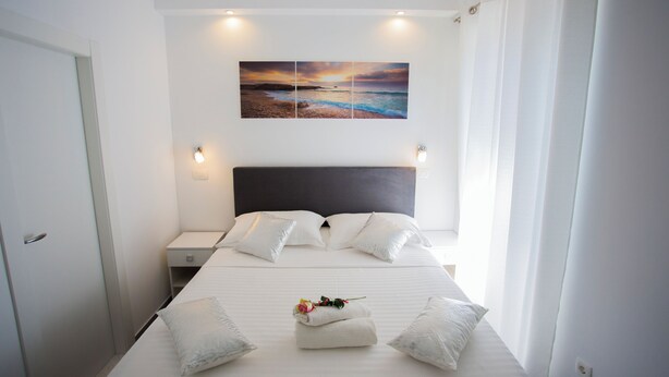 A neatly made bed with white linens and pillows, a bedside table with a lamp, and a wall-mounted painting of a beach scene. - Nirvana Garden Rooms (Split)