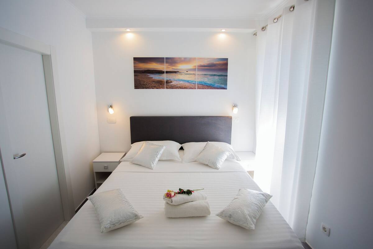 A neatly made bed with white linens and pillows, a bedside table with a lamp, and a wall-mounted painting of a beach scene.