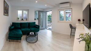 A modern living room with a green sectional sofa, a small round coffee table, and a TV mounted on the wall. There is a sliding glass door leading to an outdoor area. - Nirvana Garden Rooms (Split)