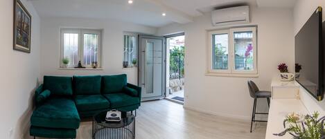 A modern living room with a green sectional sofa, a small round coffee table, and a TV mounted on the wall. There is a sliding glass door leading to an outdoor area.