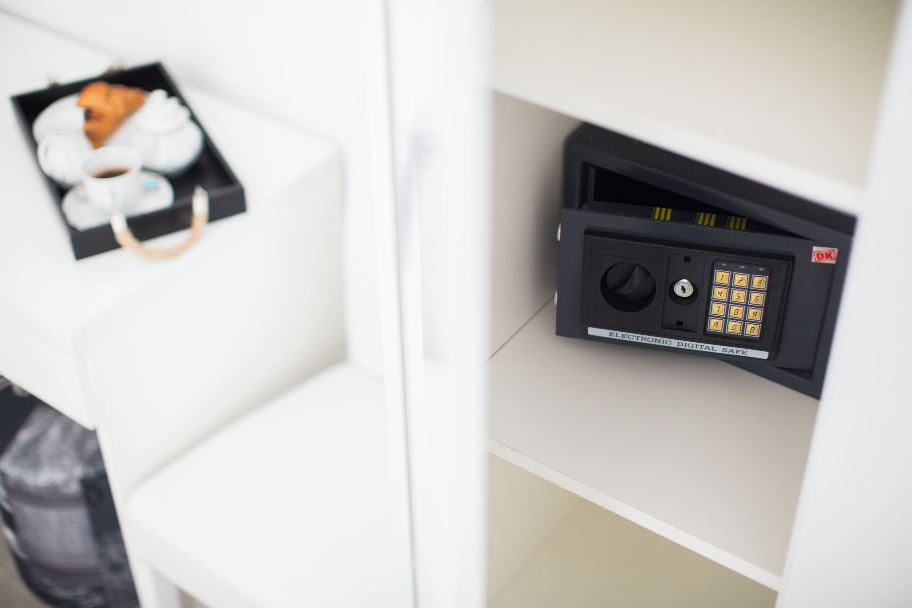 A digital safe with a keypad and a lock mechanism, a small tray with a croissant and a cup of coffee, and a black bag on a white surface.