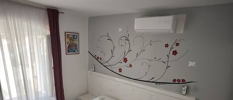 Un dormitorio con cama blanca, cabecero con diseño floral, mesita de noche y velador.