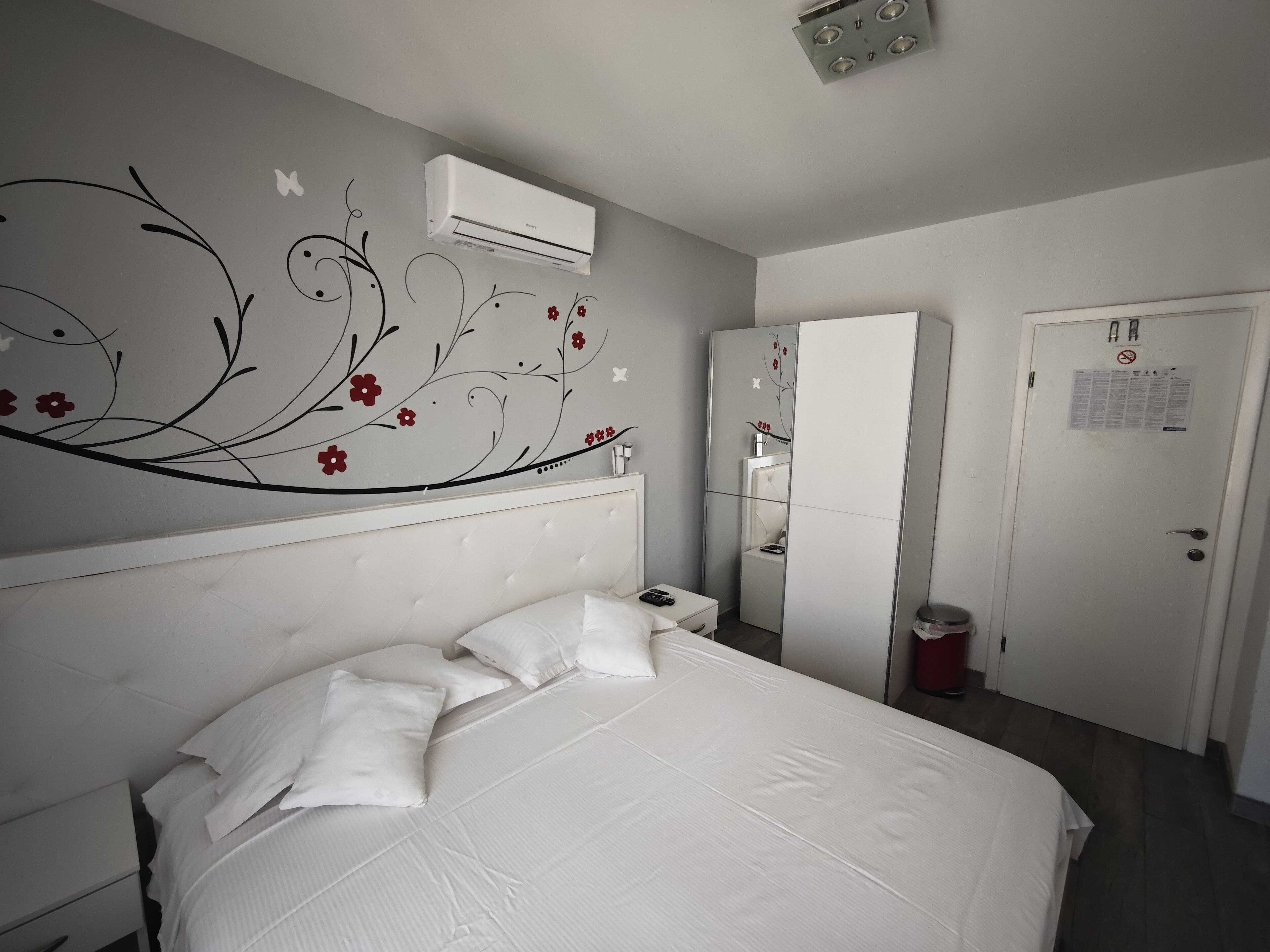A bedroom with a bed, white walls, a decorative floral wall design, a wardrobe, and an air conditioning unit.