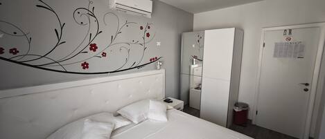 A bedroom with a bed, white walls, a decorative floral wall design, a wardrobe, and an air conditioning unit.