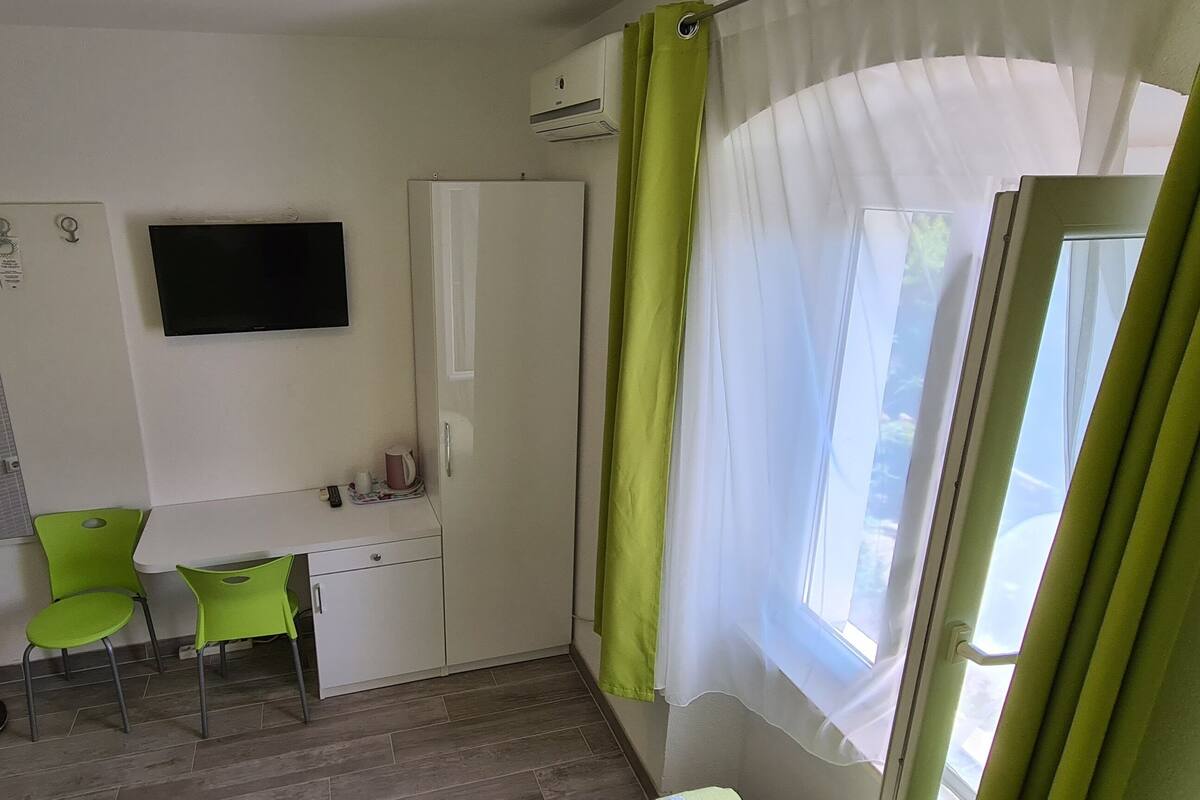 A small room with a desk, two chairs, a mirror, a TV, and a window with curtains.
