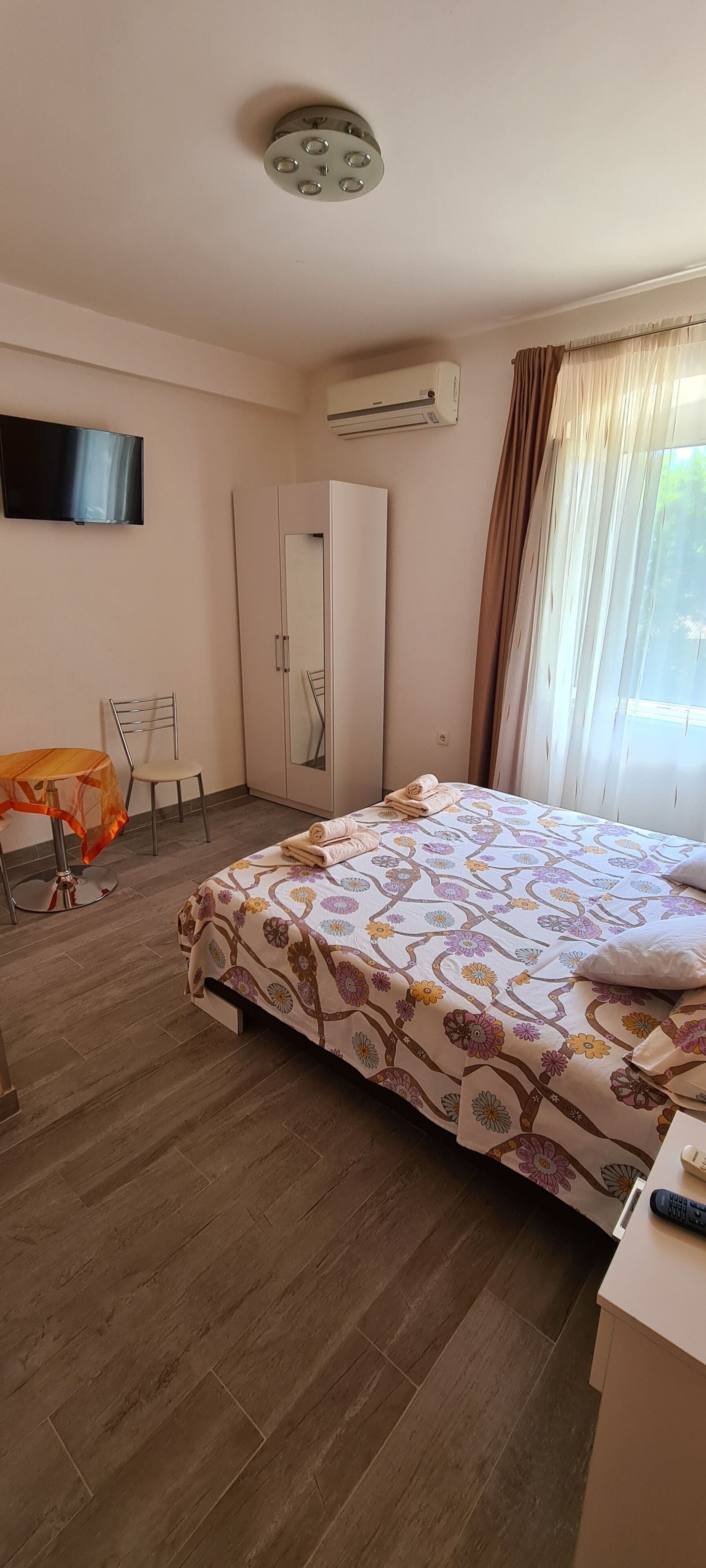 Habitación de hotel con cama, televisor, aire acondicionado, armario, silla y ventana con cortinas.