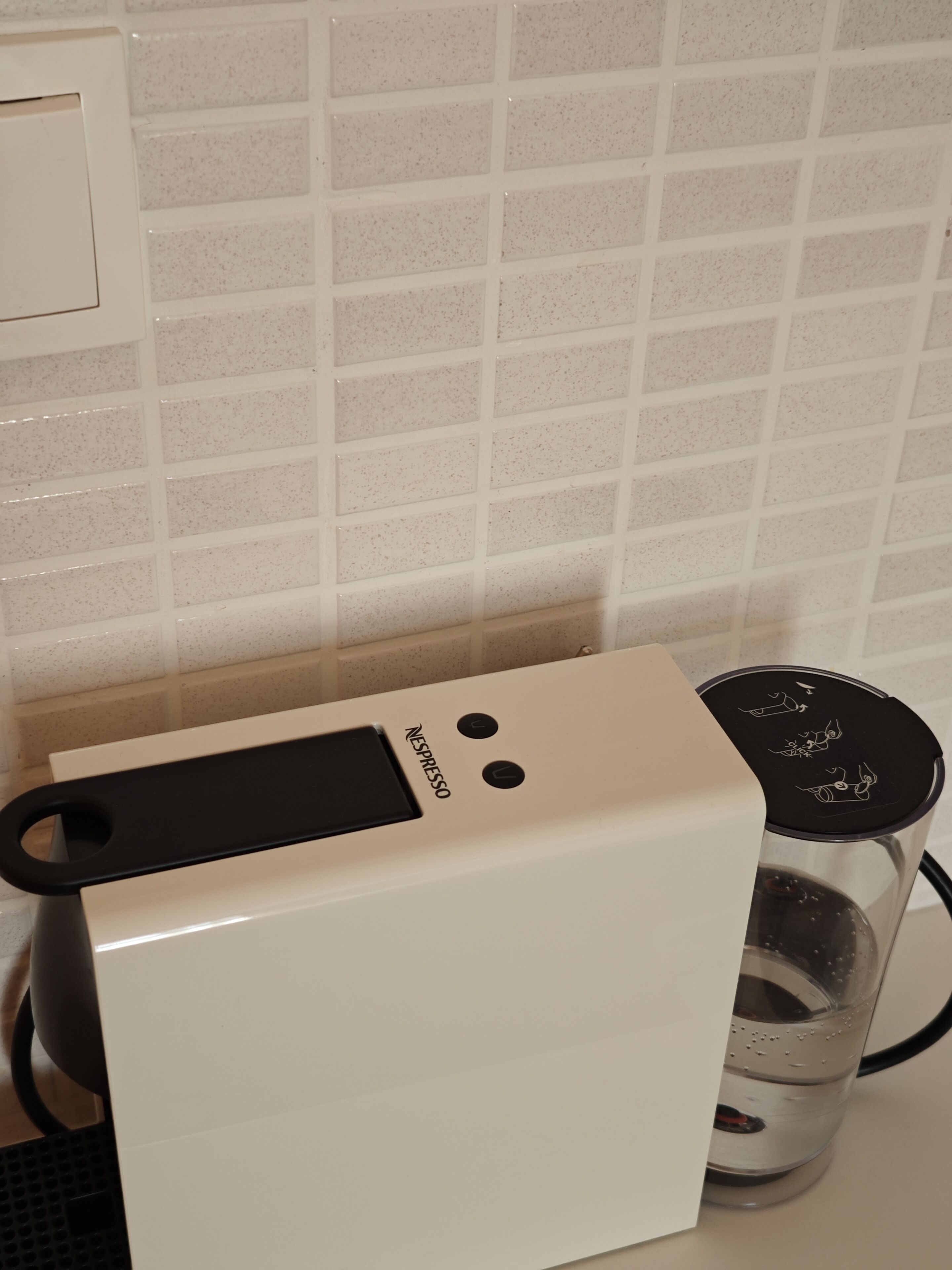 A white Nespresso coffee machine with a transparent water container beside it, set against a tiled backsplash.