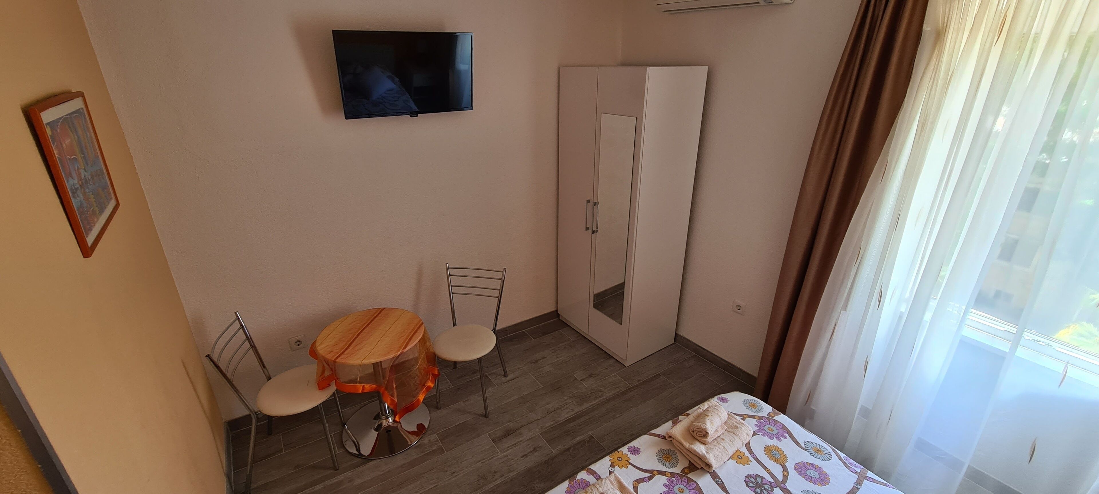 A room with a bed, a TV mounted on the wall, a wardrobe, a small round table with chairs, and a window with curtains.