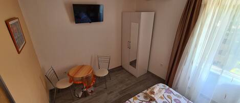 A room with a bed, a TV mounted on the wall, a wardrobe, a small round table with chairs, and a window with curtains.