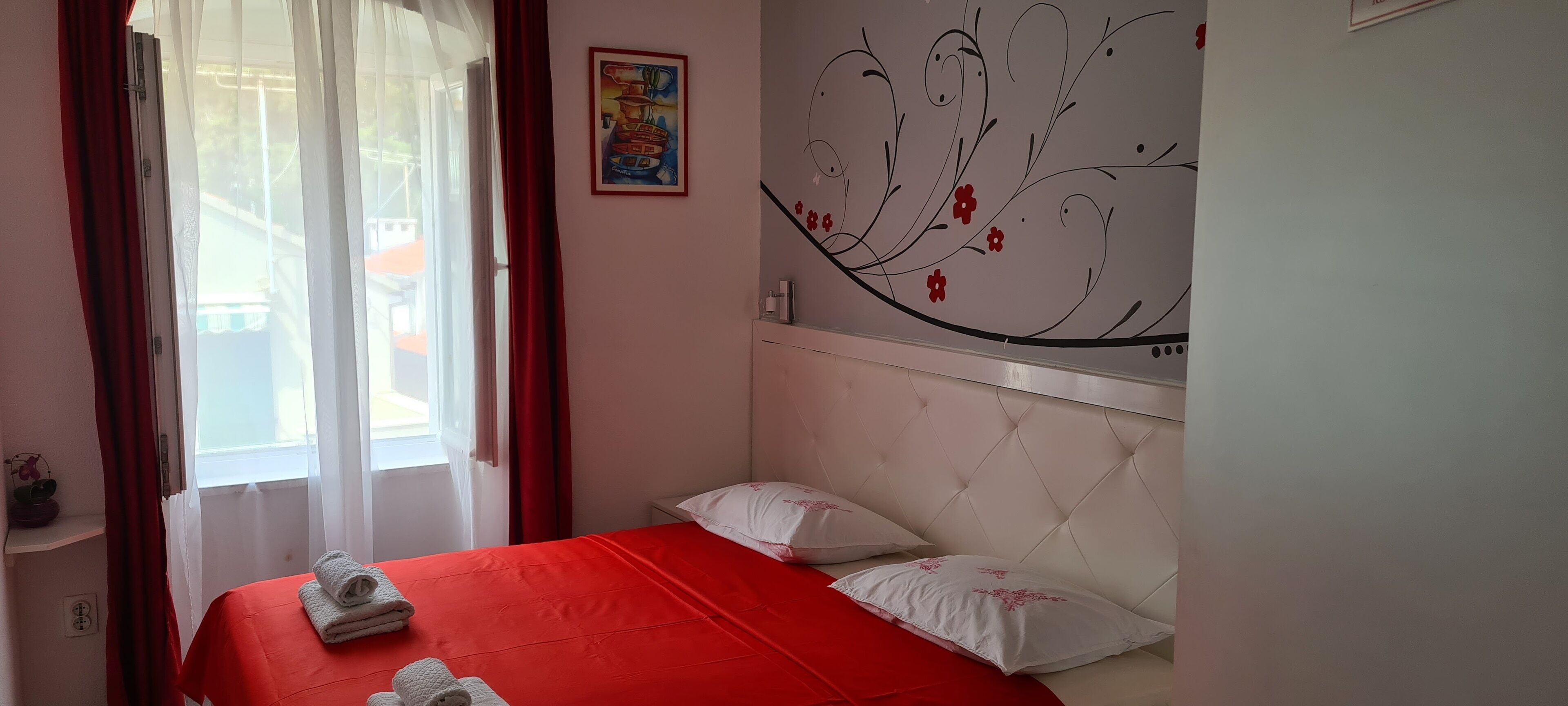 A bedroom with a red bedspread, white pillows, a decorative wall art, and a window with curtains.