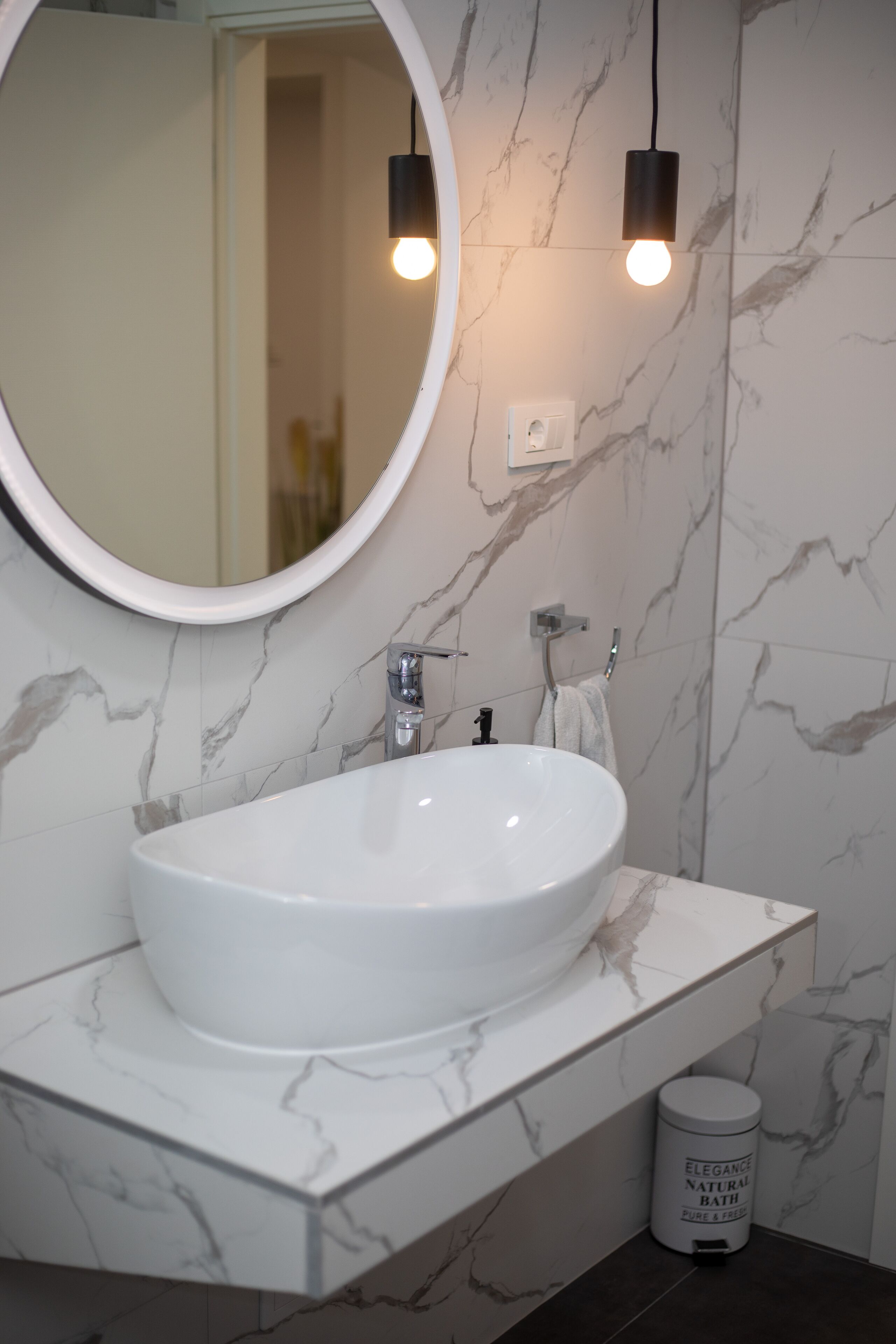 Un baño con un lavamanos blanco ovalado, un espejo redondo y dos lámparas colgantes.