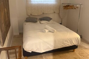 Cama bem arrumada com edredom branco, duas almofadas quadriculadas em preto e branco, e uma toalha branca enrolada.