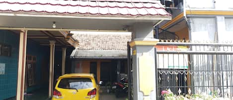 Un coche amarillo estacionado en un área de garaje techada, con techo de baldosas, una puerta metálica y un pequeño jardín con flores rosas.