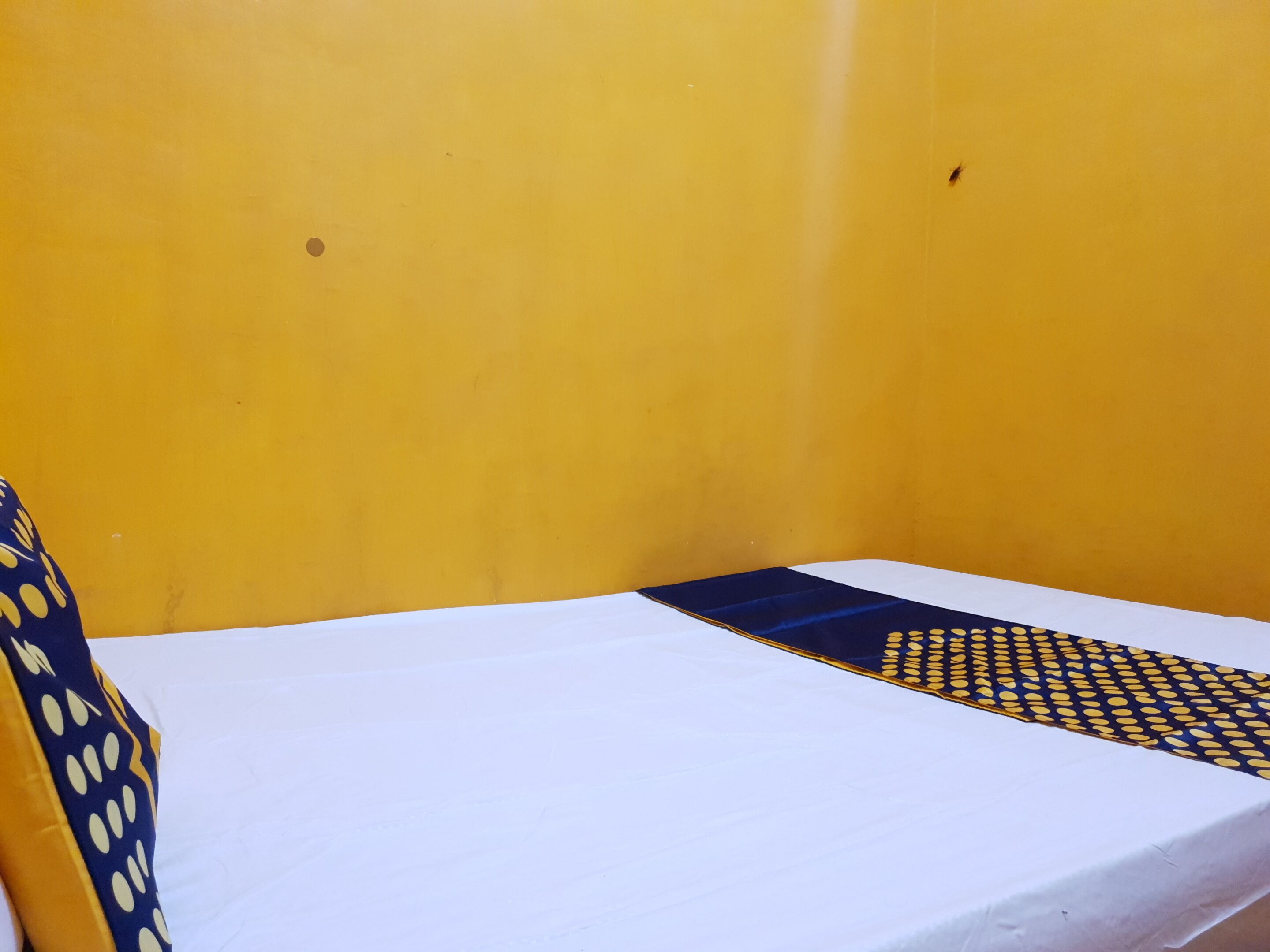 A room with a yellow wall, a white bed with a blue and yellow patterned bedspread, and a blue and yellow patterned pillow.