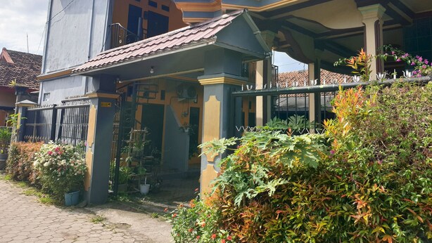 Une maison à deux étages dotée d’un balcon, d’une entrée couverte et d’un jardin où poussent diverses plantes et fleurs.
