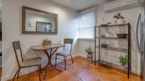 A compact kitchen with a dining table, chairs, a mirror, a window with blinds, and a metal shelf with kitchen items.