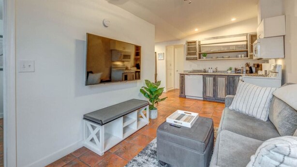 A modern living room with a grey sofa, a bench, and a coffee table. A large mirror hangs on the wall, and a ceiling fan is visible. The room leads to a kitchen with wooden cabinets and a built-in oven.