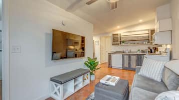 Sala de estar moderna com sofá cinza, um banco e uma mesa de centro. Um espelho grande está pendurado na parede, e um ventilador de teto é visível. O cômodo dá para uma cozinha com armários de madeira e um forno embutido.