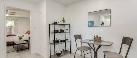 Un espacio habitable compacto con kitchenette, un área de comedor con dos sillas, una mesita y un estante metálico con ollas y sartenes.