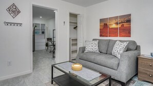 A modern living room with a grey sofa, patterned cushions, a glass coffee table, and a wall-mounted artwork depicting a sunset.