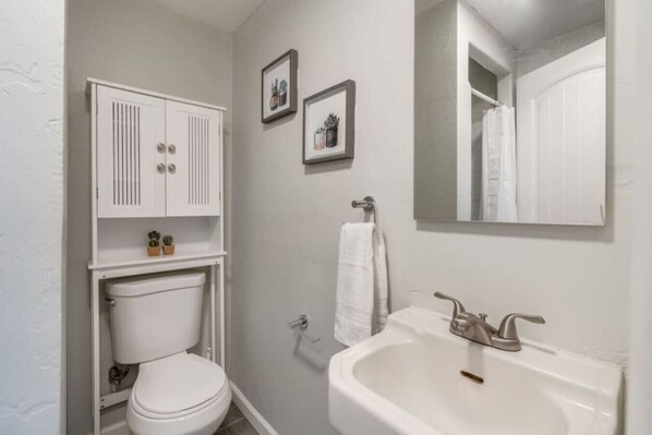 A small, white bathroom with a toilet, a sink, and a cabinet above the toilet. There are two framed pictures on the wall above the cabinet.