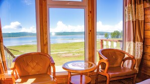 Ein Zimmer mit großem Fenster, das auf einen See und Hügel blickt. Zwei Holzstühle und ein kleiner runder Tisch sind ebenfalls vorhanden.