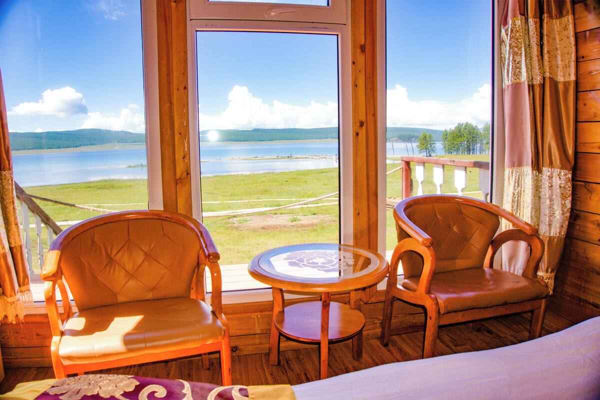 A room with a large window overlooking a lake and hills. There are two wooden chairs and a small round table.