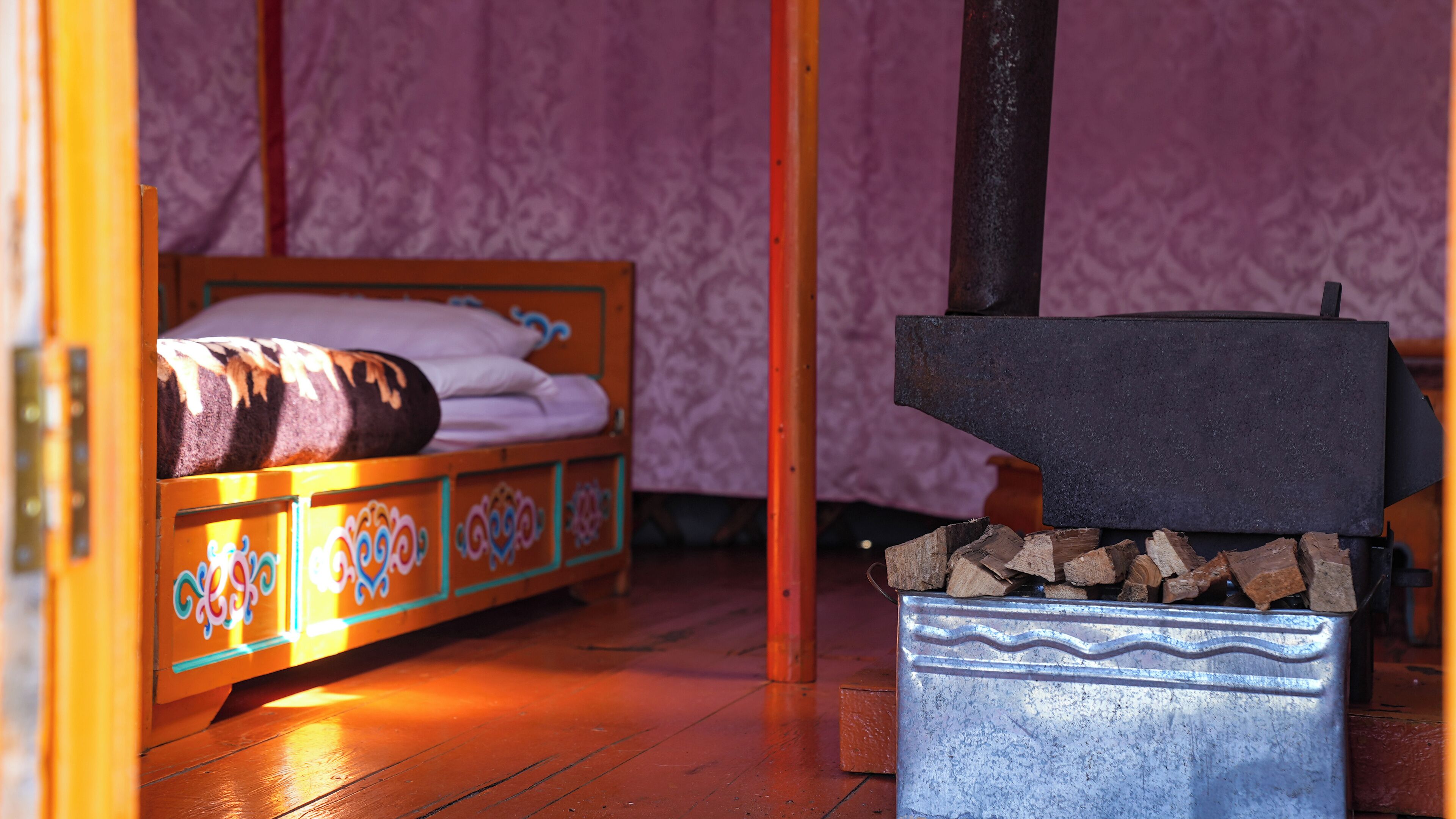 A wooden bed with colorful patterns, a metal stove with wood logs, and a purple patterned wall.