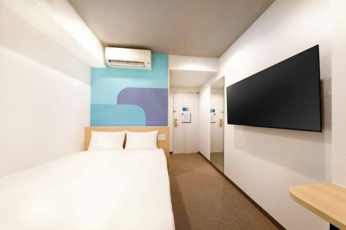 A hotel room with a bed, a TV mounted on the wall, a bedside table, and an air conditioning unit.