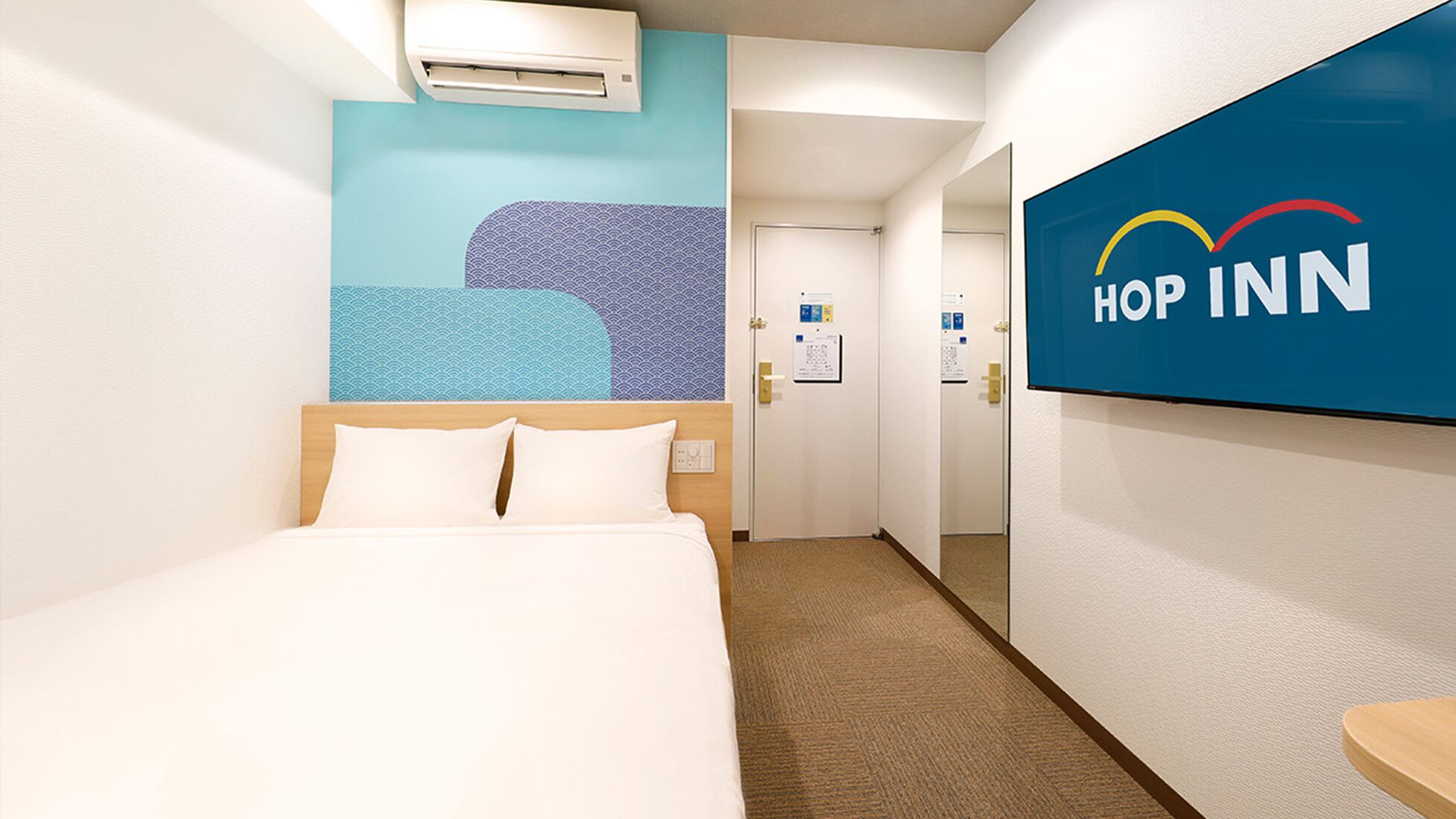 Quarto de hotel com cama, ar condicionado fixado na parede, parede azul com padrão de ondas e placa com o logotipo 'HOP INN'.
