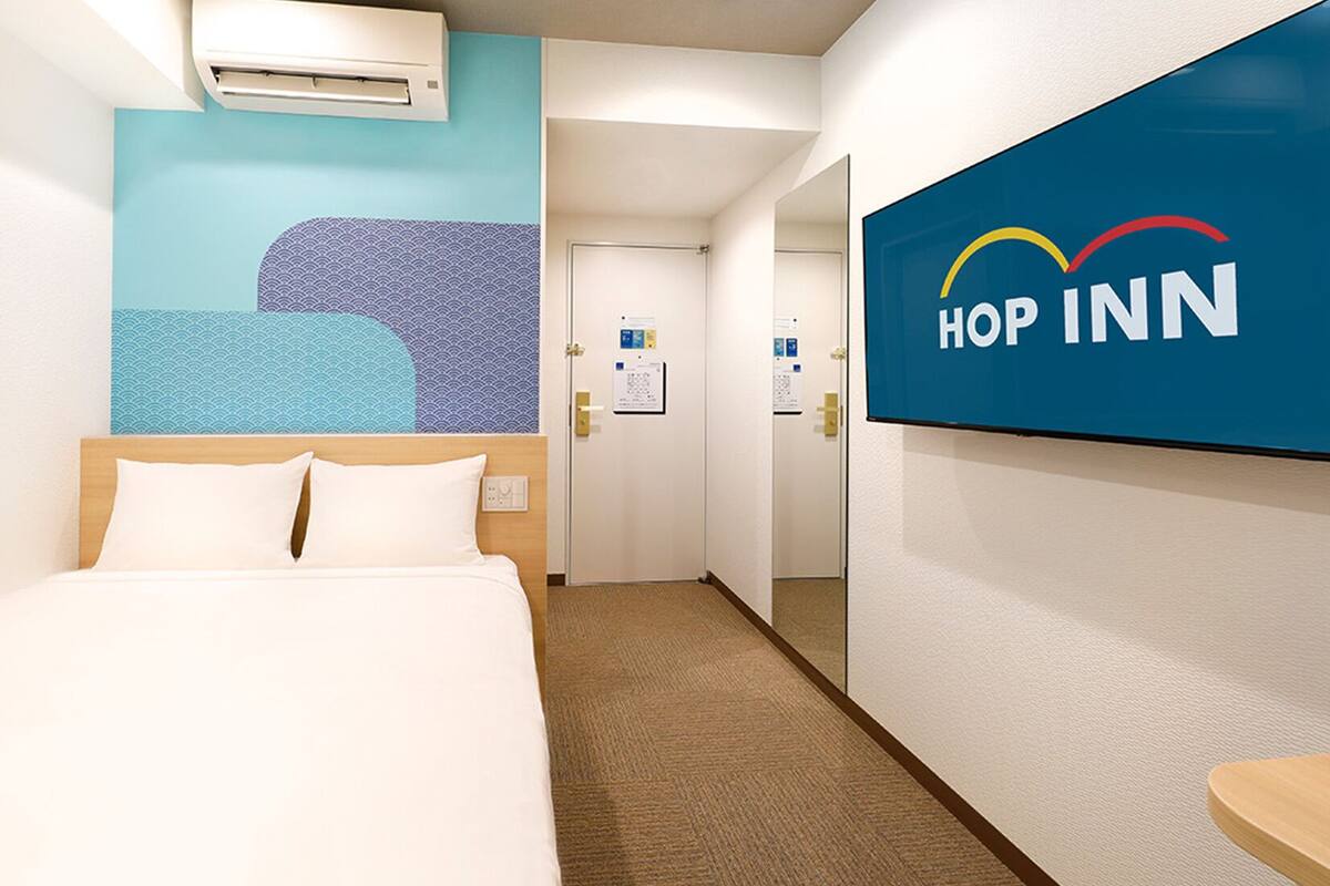 A hotel room with a bed, a wall-mounted air conditioner, a blue wall with a wave pattern, and a sign with 'HOP INN' logo.
