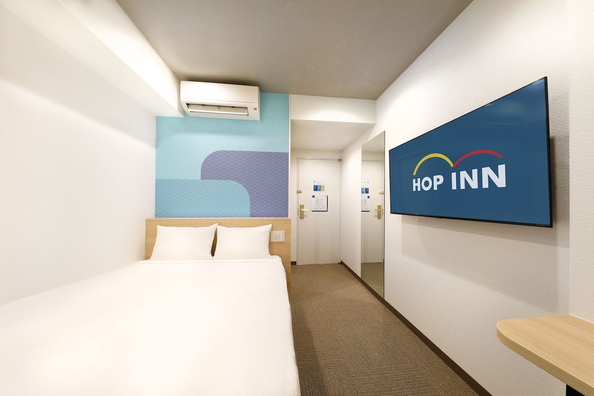 A hotel room with a bed, a wall-mounted TV displaying the 'Hop Inn' logo, an air conditioning unit, and a patterned wall design.