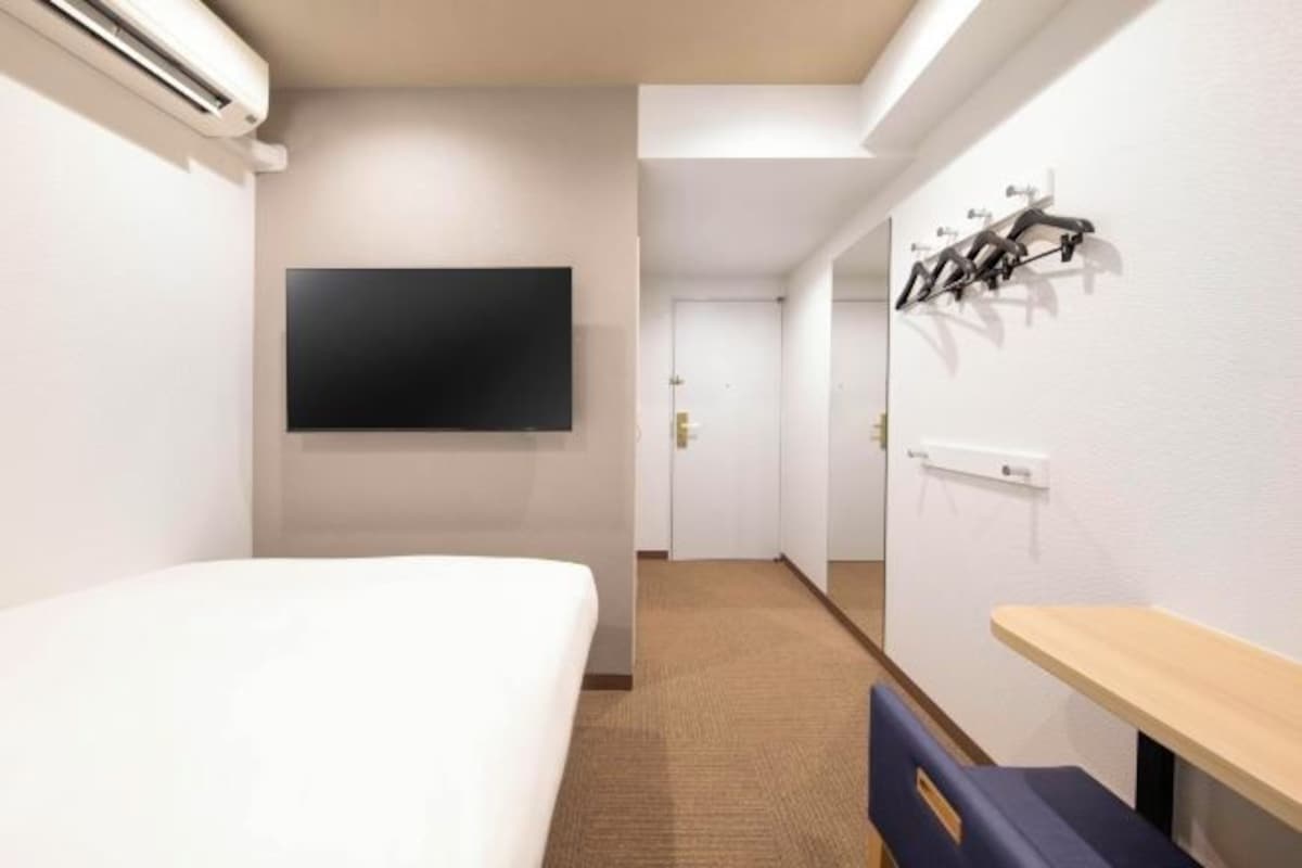 A hotel room with a bed, a flat-screen TV mounted on the wall, a coat rack, and a wooden bench.