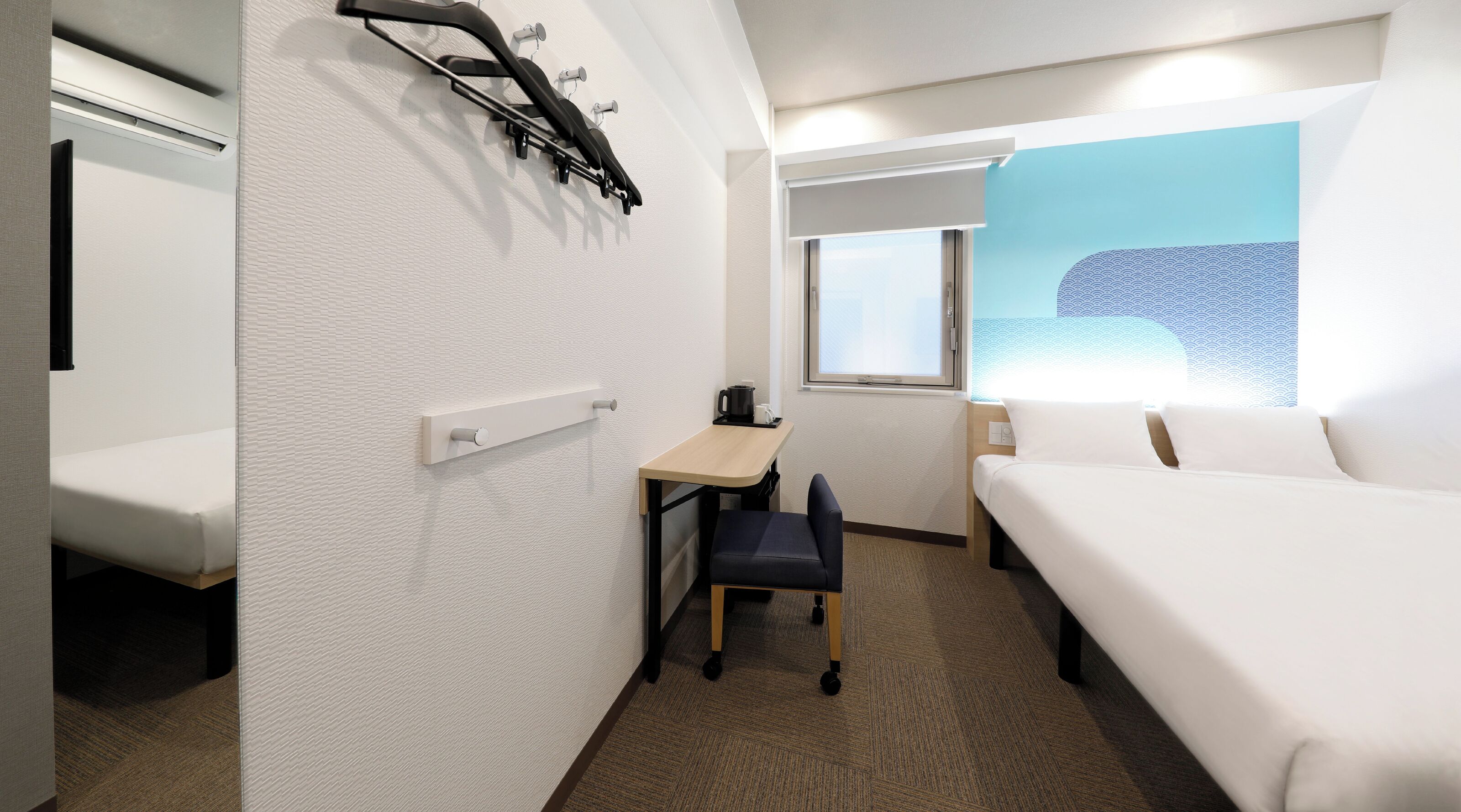 Quarto de hotel com cama, escrivaninha, cadeira, janela e aquecedor fixado na parede.