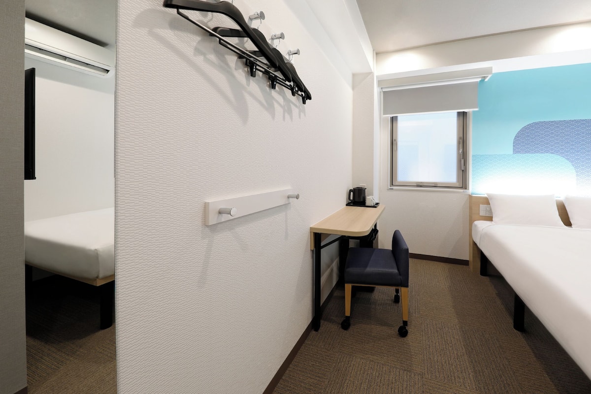 A hotel room with a bed, a desk with a chair, a window, and a wall-mounted heater.