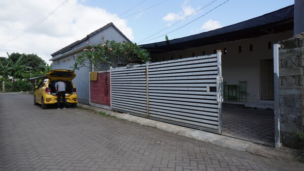 A yellow car parked in front of a gated area with a brick pathway leading to a building with a tiled roof. - Hotel O Hunian 99 (Genteng)