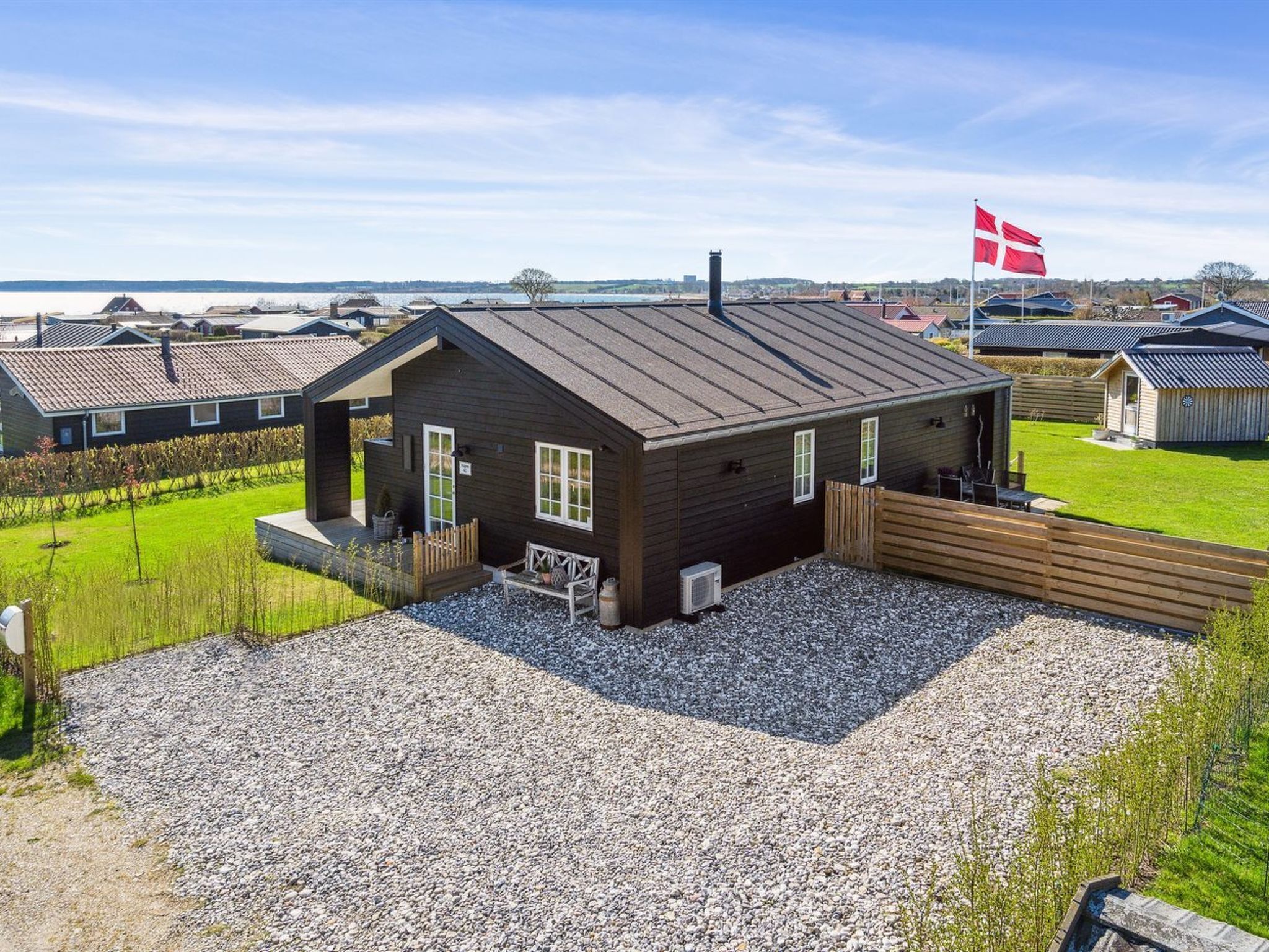 Een klein, donker houten huisje met een grindpad en een houten terras, omgeven door een grasveld en een vlag.
