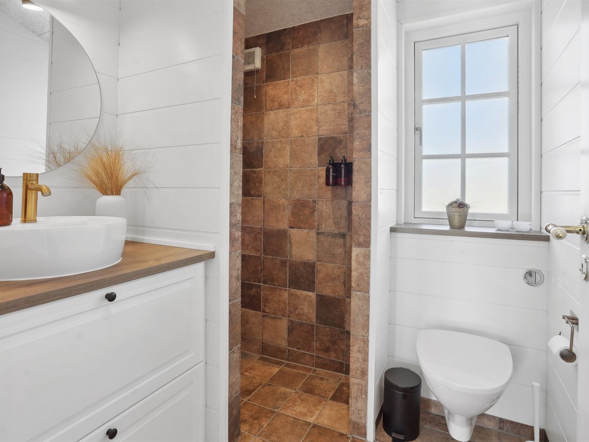 A bathroom with a white sink, gold faucet, and a window with a view.