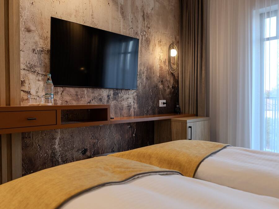 A hotel room with a large flat-screen TV mounted on a textured wall, a wooden bedside table with a bottle and glasses, and a bed with a yellow blanket.