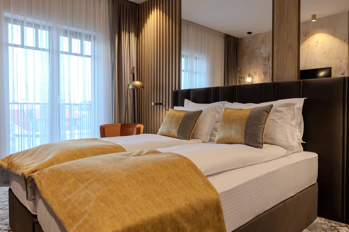 Ein Hotelzimmer mit einem großen Bett, einem Sessel, einer Lampe und Blick ins Freie durch das Fenster.