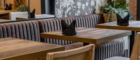 Interior de restaurante con mesas de madera y cabinas acolchadas, con un llamativo papel tapiz floral y plantas en macetas.