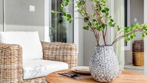 Un canapé en osier avec un coussin blanc, une table ronde en bois surmontée d’un vase texturé et d’un verre, et un panier en osier en arrière-plan.