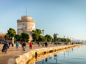 A historic tower with a circular base and a flagpole, surrounded by a promenade with trees and a water body reflecting the structure. - Sanders Old Port - Darling 1-bdr. Apt. w/ Balcony (Thessaloniki)