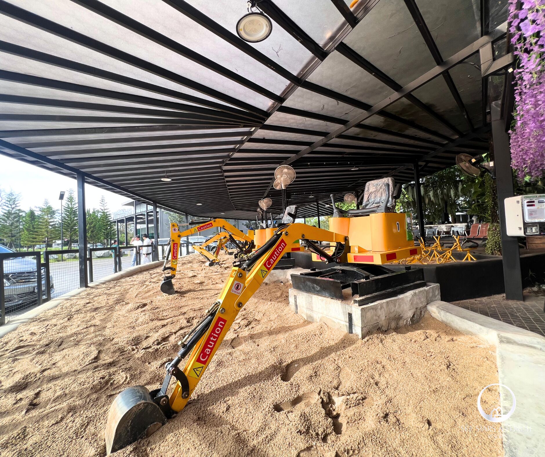 Una obra en construcción con una excavadora amarilla, un área techada con techo metálico y una estructura de concreto.
