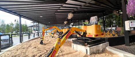 Una obra en construcción con una excavadora amarilla, un área techada con techo metálico y una estructura de concreto.
