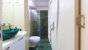 Ein schmaler Flur, der zu einem Badezimmer mit einem grünen Waschbecken, einer Toilette und einer Duschkabine aus Glas führt.